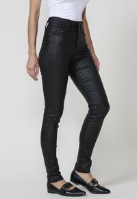 Crne visoke hlače s teksturiranim uzorkom houndstooth, uskog kroja i prednjim džepovima, uparene s ravnim crnim cipelama s metalnim detaljima.