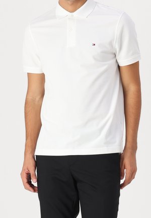 Mann trägt ein schlichtes weißes kurzärmeliges Poloshirt mit kleinem Logo auf der linken Brust und schwarze Hose vor weißem Hintergrund.