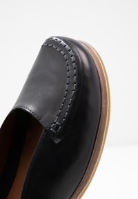 Zapato negro de cuero sin cordones con detalles de costura, punta redondeada y suela de goma marrón claro. Textura suave con un acabado pulido.