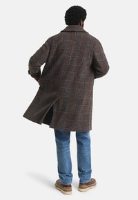 Man met krullend haar, gekleed in een bruine geruite jas, blauwe jeans en beige schoenen, staande met zijn rug naar voren. 