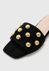 Black suede slide sandal featuring a wide strap adorned with gold metallic studs. Square toe design with a flat sole.