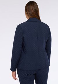 Námořnický modrý blazer s přizpůsobeným střihem, s texturovaným povrchem a malými kovovými tečkami pro dodatečný detail, pohled ze zad.