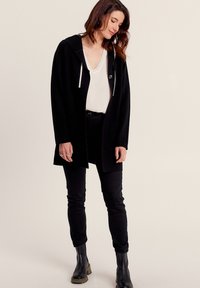 Cappotto nero con cappuccio, realizzato in tessuto morbido, dotato di lacci bianchi, tasche frontali, abbinato a una blusa crema e pantaloni neri.