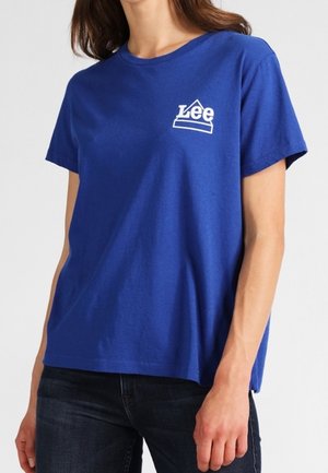 Kobieta ubrana w niebieski krótki t-shirt Lee z białym logo na piersi oraz ciemne jeansy, stojąca na jasnym, jednolitym tle.