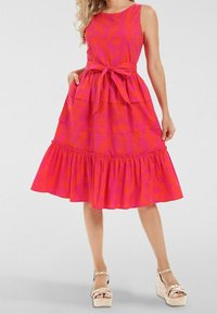 Robe A-line rose avec motif floral, design sans manches, ceinture à la taille, jupe superposée et ourlet volanté. Associée avec des sandales compensées beiges.