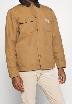 Homme portant une veste beige à boutons avec quatre poches avant et un pantalon beige clair, debout devant un fond uni.