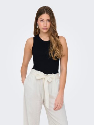 Joven mujer con cabello largo y ondulado lleva una camiseta negra sin mangas y pantalones blancos de talle alto atados con un lazo, de pie con una mano en el bolsillo.