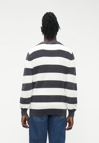 Oseba z rasta pričesko, oblečena v sivo-bel pulover s črtami in modre kavbojke, obrnjena stran, na svetlem enobarvnem ozadju.