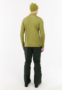 Man in olijfgroen overhemd met lange mouwen, bijpassende beanie, donkergroene broek en zwarte laarzen, staand met zijn rug naar de camera tegen een witte achtergrond.