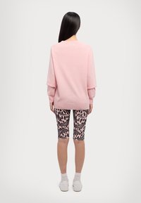 Pull rose clair à coupe ample et manches longues, assorti à un short de vélo noir et rose à motifs. Le modèle porte des baskets blanches.