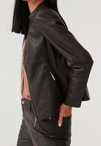 Veste en cuir brun foncé avec une coupe slim, col ouvert et poches latérales zippées. Texture douce et détails de design minimalistes.