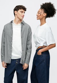 Ριγέ πουκάμισο με κουμπιά πάνω από ένα λευκό T-shirt· μοντέλο με σκούρα μπλε παντελόνια φαρδιάς γραμμής. Ένα άλλο μοντέλο με λευκό T-shirt δεμένο κόμπο και ψηλόμεσα τζιν.