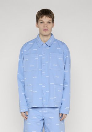 Mladić u svijetloplavoj jakni na kopčanje i hlačama s ponavljajućim uzorkom logotipa "ecko unltd.", stoji ispred jednostavne pozadine.