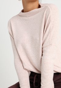 Heller rosa, strukturierter Pullover mit Boatneck und überschnittenen Schultern. Weicher Stoff, lässige Passform. Modell trägt im Vordergrund dunkle Hosen.