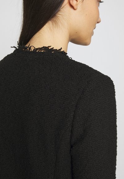 Veste noire texturée avec un accent effiloché au niveau du col. Présente une silhouette structurée et un tissu doux et tissé.