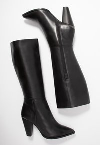Par de botas de cuero negro hasta la rodilla con puntas afiladas, tacones de bloque y cremalleras laterales sobre fondo blanco.