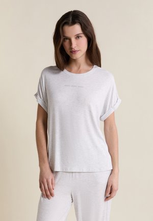 Jeune femme aux cheveux bruns raides portant un t-shirt gris clair à manches retroussées et un pantalon assorti, debout devant un fond beige.