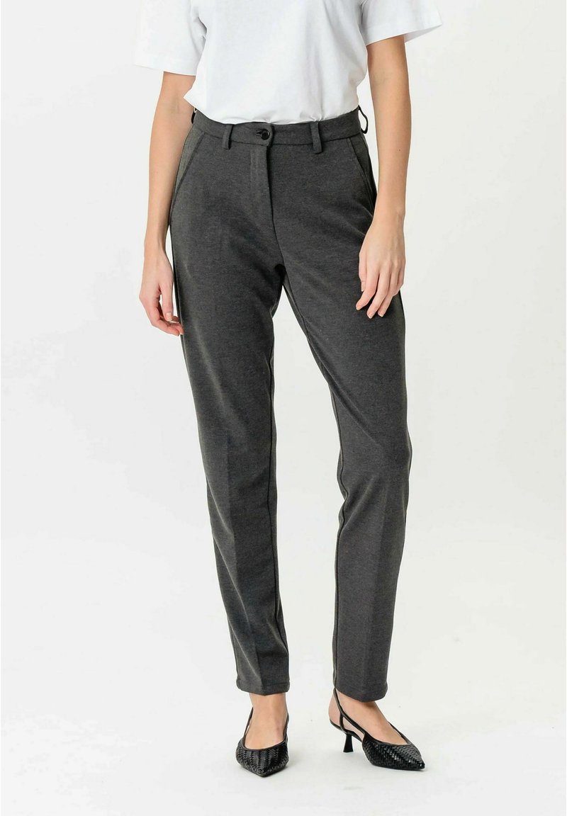 Femeie purtând pantaloni conici gri închis cu buzunare laterale și închidere cu nasture, asortați cu pantofi negri cu toc mic și bretea la spate.