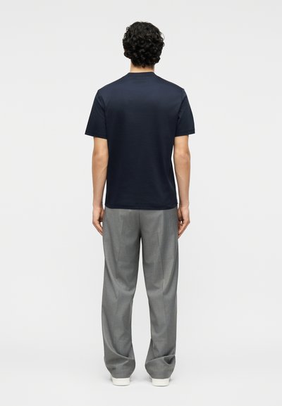 Άνδρας με σγουρά σκούρα μαλλιά στέκεται με την πλάτη, φοράει ναυτικό μπλε κοντομάνικο πουκάμισο, γκρι παντελόνι και λευκά παπούτσια μπροστά σε απλό φόντο.