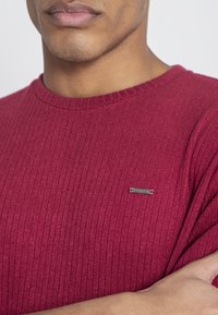 Röd ribbstickad tröja med rund halsringning och en liten metallisk logotyp på bröstet. Tyget verkar mjukt och texturerat.