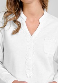 Mujer con una camisa blanca de manga larga, con encaje en los bordes, botones en el frente, un pequeño bolsillo y un lazo de satén en la parte inferior.