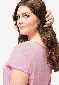 Top rosa chiaro a maniche corte con scollatura arrotondata e dettaglio intrecciato sul retro. Tessuto morbido con vestibilità comoda, che mostra una leggera caduta.