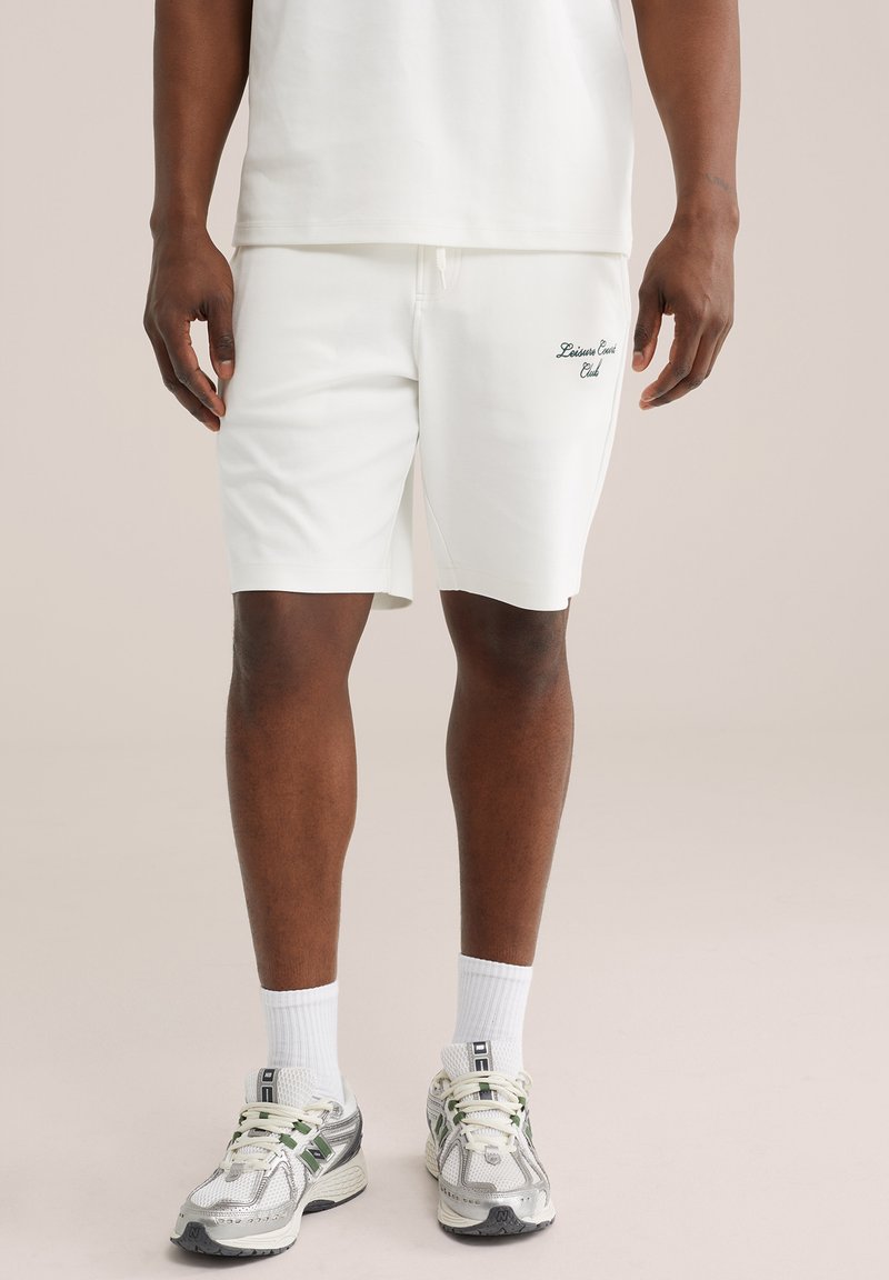 Homme portant un short blanc avec le texte « Leisure Court Club », des chaussettes blanches et des chaussures de course blanc-gris, debout sur un fond neutre.