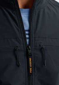 Giacca blu scuro con zip completa, due tasche frontali con cordini e un'etichetta con logo sulla zip. Sotto, una camicia azzurra chiara.