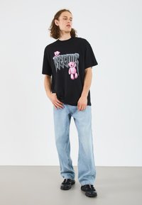 T-shirt nera con una grafica di un orsetto rosa e testo, abbinata a jeans larghi azzurri chiari e sneakers nere.