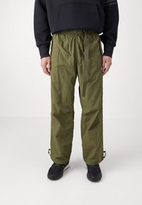 Olivgröna, lätta byxor med avslappnad passform, elastisk midja och justerbart bälte. Har dragkedjor vid anklarna och en slät textur.