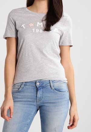 Kobieta ubrana w jasnoszarą koszulkę z napisem "TOMMY 1985" i grafiką gwiazdy, połączoną z jasnoniebieskimi, wyblakłymi jeansami.