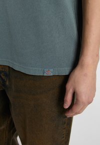 Camiseta gris de manga corta hecha de algodón con un acabado ligeramente texturizado; cuenta con una pequeña etiqueta de logo colorido de Dickies en el dobladillo.