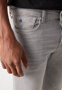Grijze denim jeans hebben een voorzak met zichtbare stiksels en een klein blauw logo. De hand rust nabij het bovenbeen.