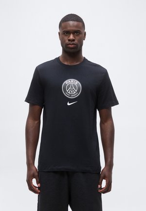 Mężczyzna stoi, ubrany w czarną koszulkę Nike Paris Saint-Germain i ciemne spodnie na jednolitym jasnym tle.