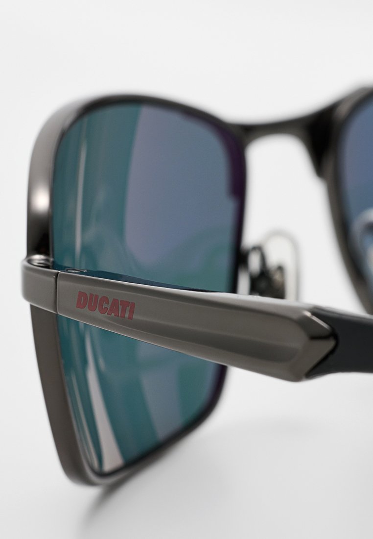 Les lunettes de soleil Ducati possèdent une monture sombre, un design rectangulaire et des verres bleu iridescent. La branche affiche un logo rouge "DUCATI".