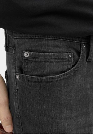 Gros plan sur la poche d'un jean en denim noir avec des rivets métalliques, porté avec une chemise noire partiellement visible au-dessus de la ceinture.