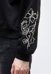 Μανίκι μαύρου φούτερ με λευκή κεντητική με λουλούδια, φορεμένο από ένα άτομο με σκούρο τζιν, το χέρι χαλαρό στο πλάι μπροστά από απλό φόντο.