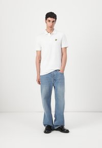 Vit polo med en gul fjärilslogga, kombinerad med ljusblå vida jeans och svarta skor. Enfärgade med minimala detaljer.