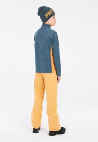 Fleece-pullover i teal med orange sidepaneler, parret med orange bukser og en hue med iøjnefaldende "PRO" tekst. Sorte fodtøj.