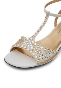 Beige Sandale mit T-Riemen Design, verziert mit goldenen und klaren Strasssteinen. Glatte Lederinnensohle mit flacher schwarzer Außensohle.