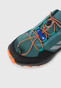 Türkisfarbene Mesh-Laufschuhe mit schwarzer und oranger Gummisohle. Mit orangenen Schnürsenkeln, blauen Akzenten und reflektierendem silbernen Logo. Die Textur ist atmungsaktiv.