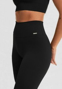 Black ribbed leggings with a high waist, featuring a metallic logo accent. Made of stretchy material, emphasizing a fitted design.