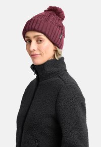 Cappello di lana marrone con un pompon sopra, caratterizzato da un motivo testurizzato. Abbinato a una giacca in pile nera, che mostra una consistenza morbida e pelosa.