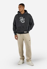 Gray pullover hoodie with a large graphic logo, paired with beige ribbed pants and white sneakers; features drawstring hood and front pocket.