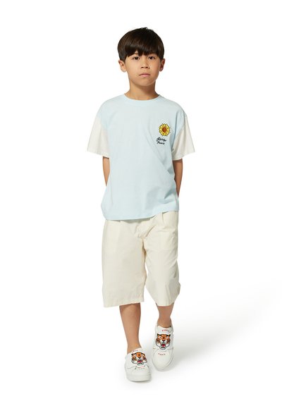 T-shirt bleu clair à manches courtes avec des manches blanches et une broderie de tournesol ; shorts crème ; chaussures blanches avec un motif de tigre.