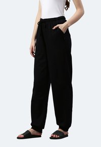 Schwarze Jogginghosen mit elastischem Bund, Kordelzug, Seitentaschen und schmal zulaufenden Bündchen. Kombiniert mit schwarzen Rutschsandalen.