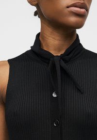 Γυναίκα με μαύρο αμάνικο ριμπ τοπ με κουμπιά στο μπροστινό μέρος και λεπτομέρεια δεσίματος στο γιακά, εμφανίζεται από τους ώμους μέχρι το πηγούνι.