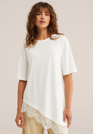 Kobieta o falowanych włosach, ubrana w asymetryczny biały top z krótkim rękawem i koronkową lamówką, stojąca na tle o neutralnym kolorze, w beżowych spodniach.