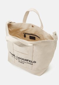 Borsa tote in canvas beige con testo stampato nero. Presenta due manici robusti, una tasca interna con zip e un design ampio e aperto.