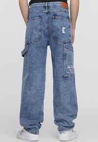 Light blue denim cargo pants with a loose fit, multiple pockets, and a logo patch on the rear, paired with white sneakers.